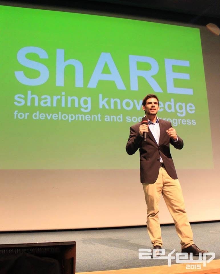 Francisco on stage with a microphone, presenting in front of a ShARE slide